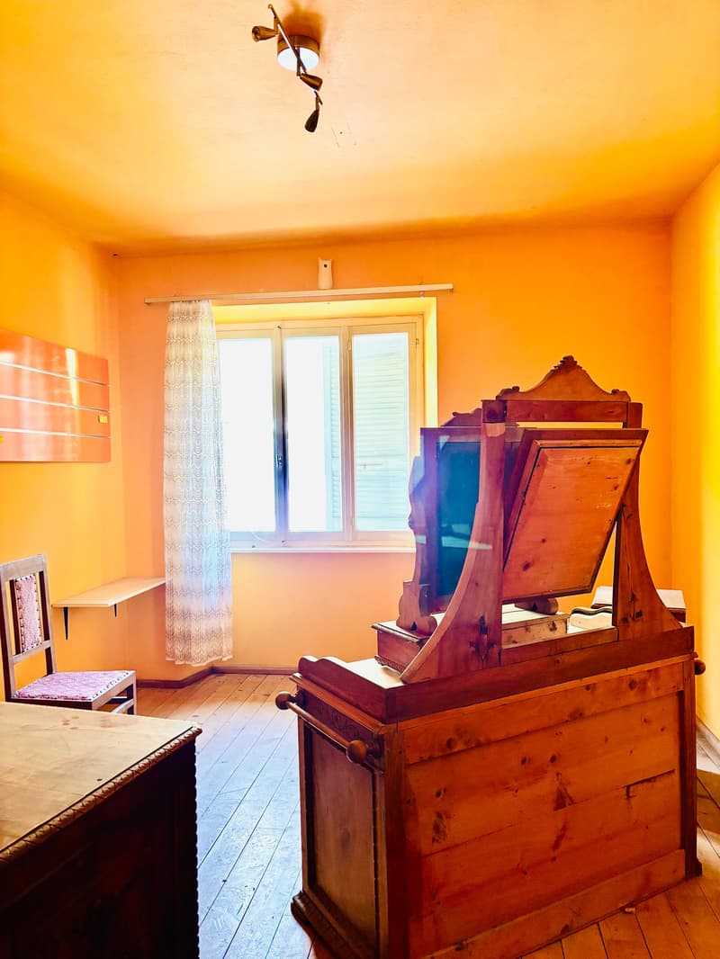 The image shows a cozy room with warm, orange-yellow walls. The room features a large, antique wooden dresser or cabinet with ornate detailing. There is a window with curtains, and a wooden floor. The room has a rustic, vintage feel with the old furniture