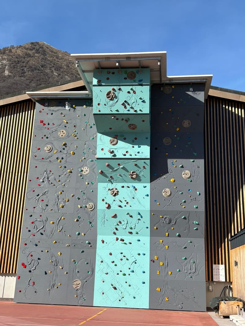 A building with a large climbing wall, different colored sections, a clear blue sky in the background, mountains in the distance.
