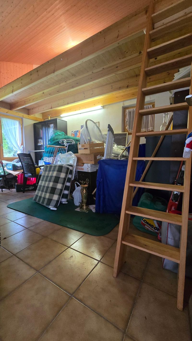 Spacious room with wooden ceiling, multiple items like crates, chair, desk, cabinet, and storage boxes, green carpet