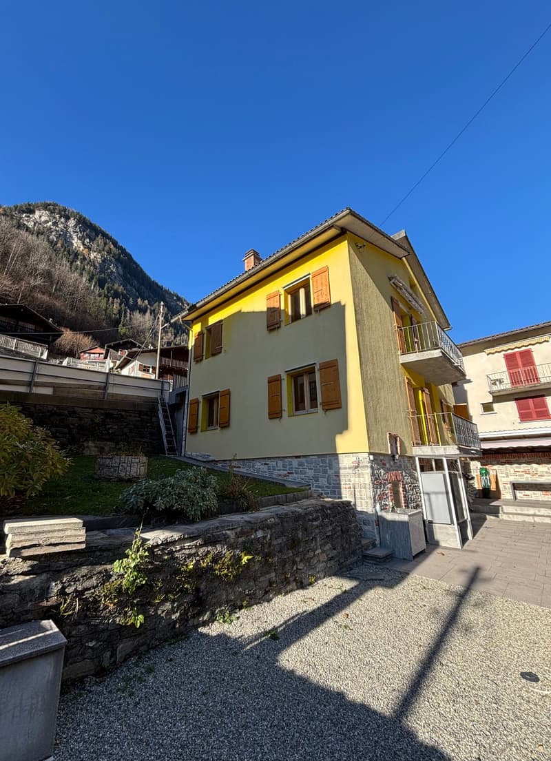 Two-story yellow house, balconies, stone base, greenery, gravel driveway, mountain view
