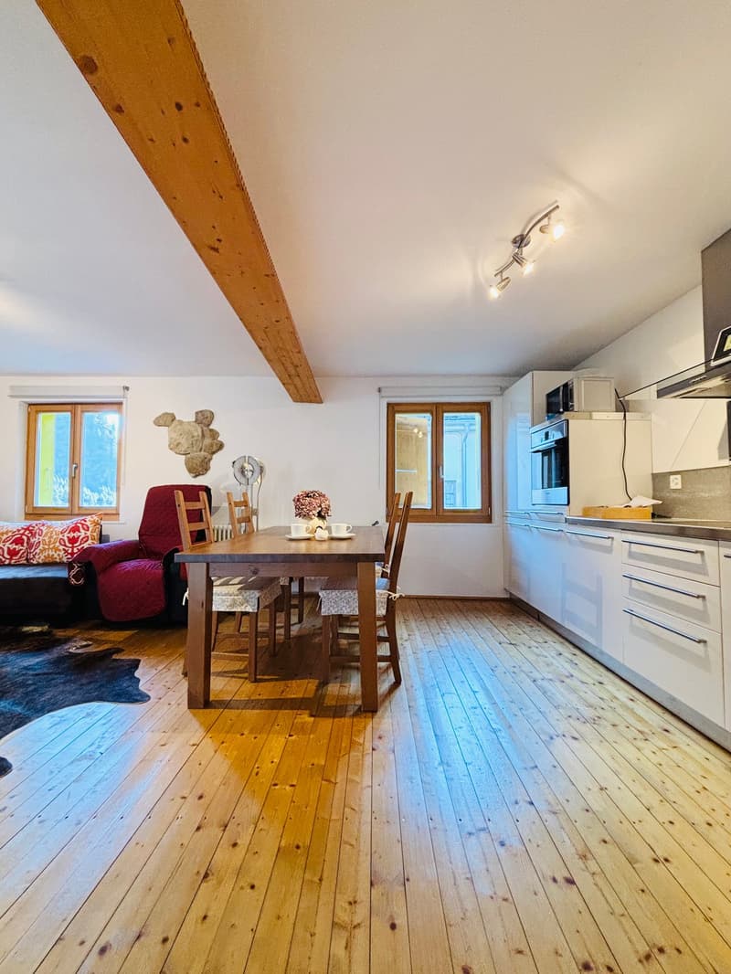 Dining room with wooden floor, dining table and chairs, kitchen with white cabinets, window, couch with pillows, radiator