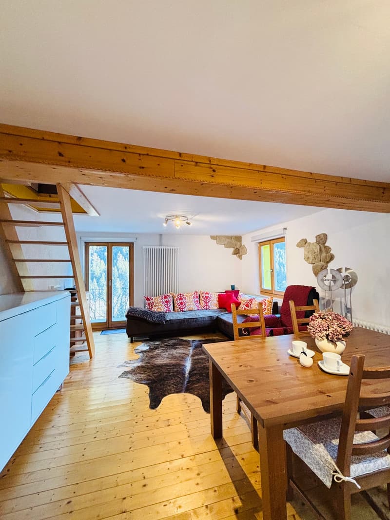 The room appears to be a living room with wooden floors, a wooden dining table with a flower vase, wooden chairs, a couch with pillows, a radiator, a staircase, and a glass door.