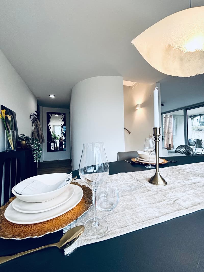 Dining room set up with a candle, plates, wine glasses, and cutlery. It has a modern, minimalist style with white walls, wooden flooring, and large windows.