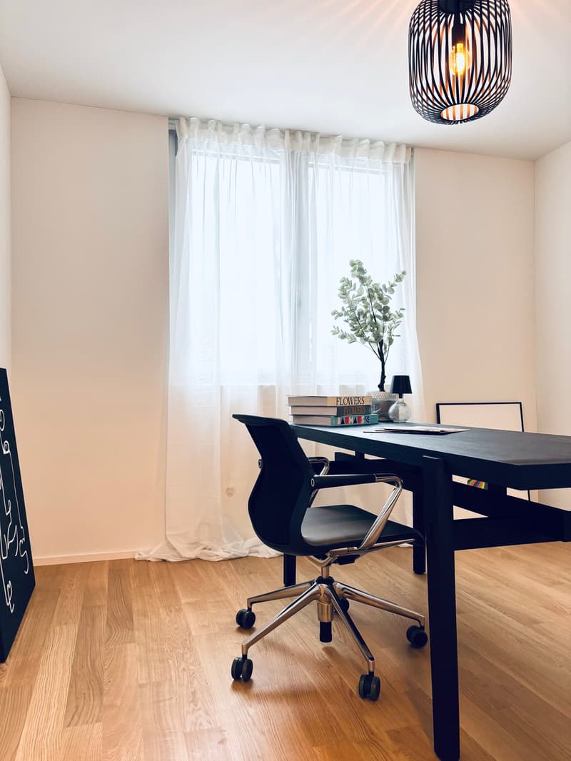 room with wooden floor, white walls, window with white curtains, office desk with rolling chair, table lamp, potted plant, books