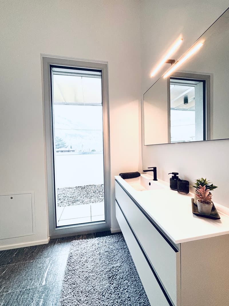 Modern bathroom with large window, white sink, black faucet, round mirror, wall mounted lighting, potted plant, and carpet