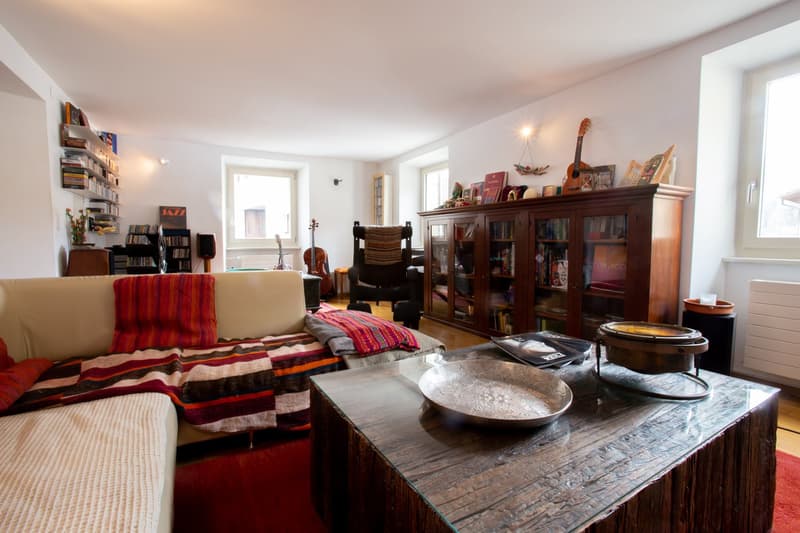 Cozy living room with hardwood floors, white walls, large windows, bookshelves, a wooden cabinet, a sofa with colorful striped cushions, a wooden coffee table with decorative bowls, and a guitar on the wall.