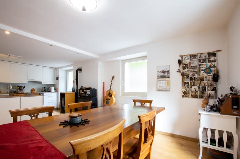 Spacious open-plan living area with a kitchen, dining table, and living room furniture. The room has large windows, hardwood floors, and various personal items and decor such as a guitar, photos, and a calendar. The space appears to be bright and airy.