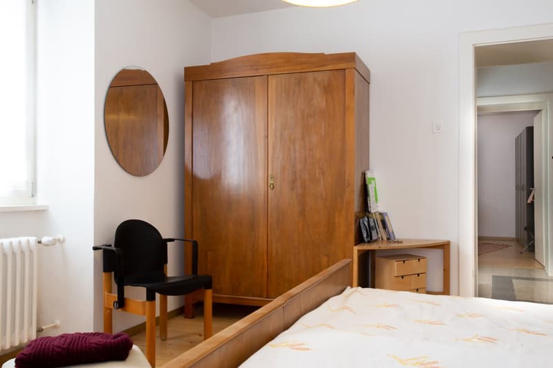 Spacious bedroom with a large wooden wardrobe, a round mirror, a black armchair, and a bed with a patterned bedspread. There is also a small side table with some books and a drawer unit.