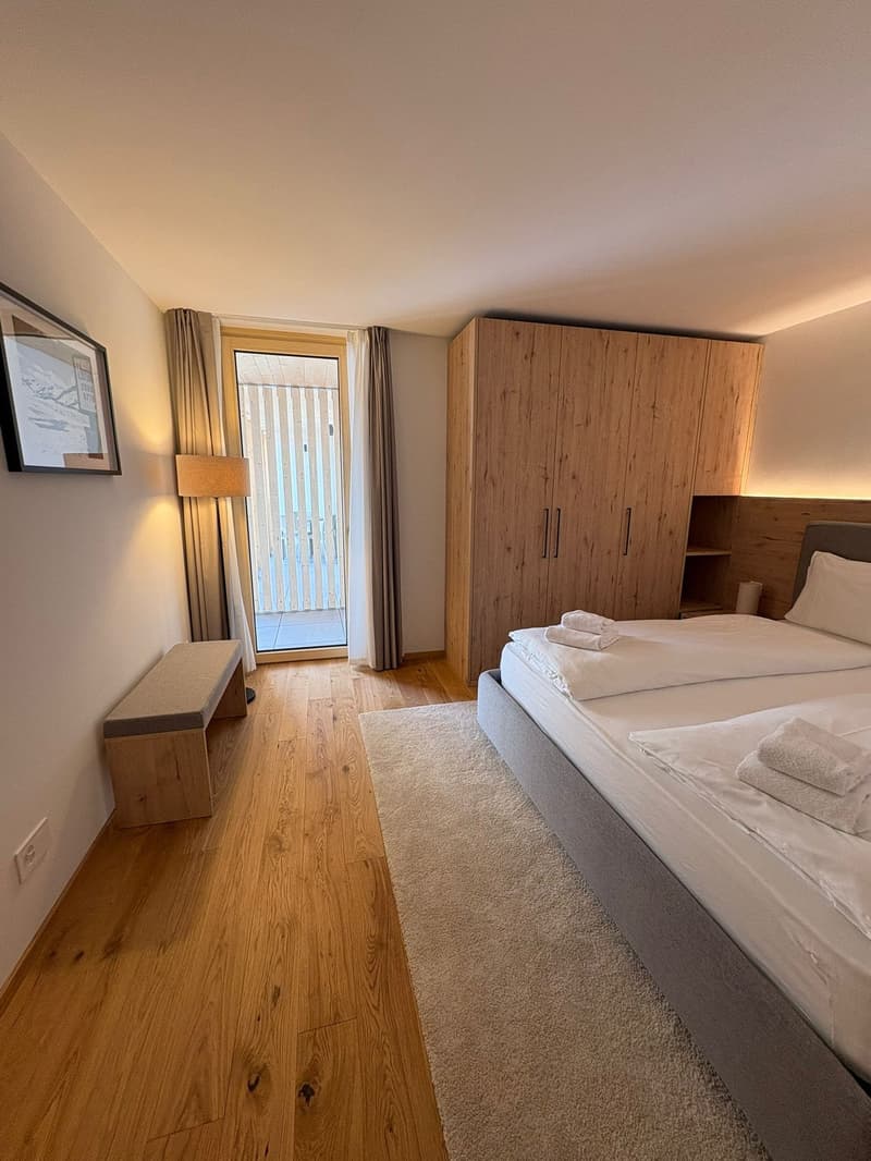 double bed, 2 pillows, towels, wooden bench, wooden closet, framed picture, lamp, power outlet, window with balcony, wooden floor