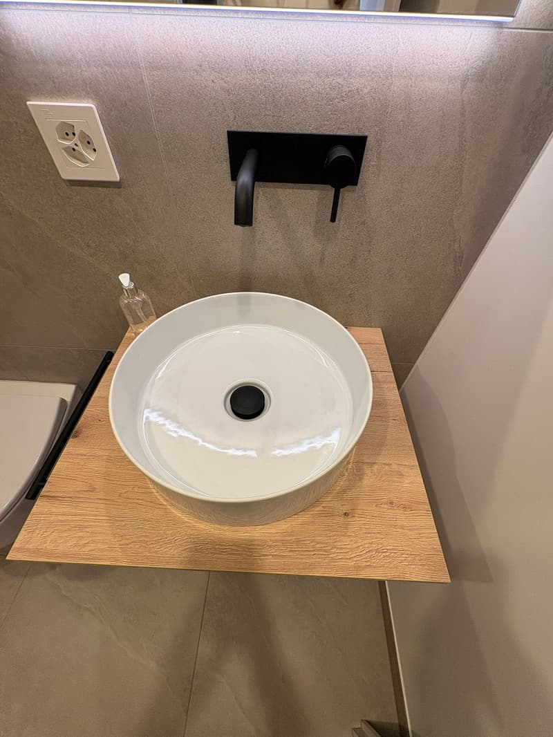 Bathroom with white basin on wooden pedestal, black tap, tiled walls, mirror, toilet, electrical outlet