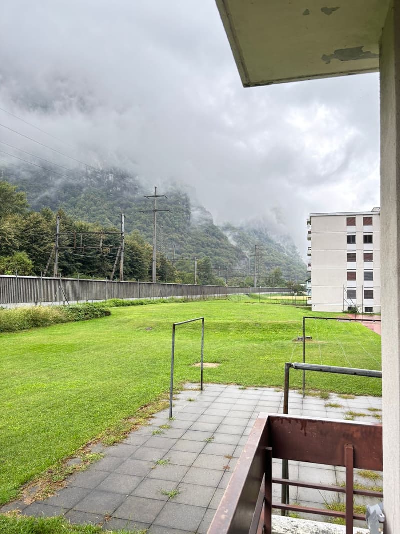 Balcony with railings, paved walkway, large grassy field, metal poles, wooden fence, distant building, mountain range, cloudy sky