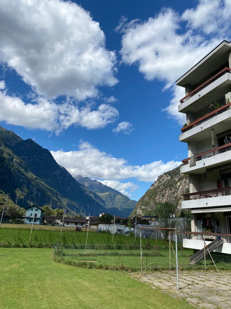A multi-story building with balconies, surrounded by a grassy field and mountains in the background