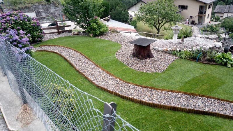 A well-maintained garden with a gravel pathway, green lawn, and various plants and flowers. There is a tree stump table and a stone lantern in the garden.