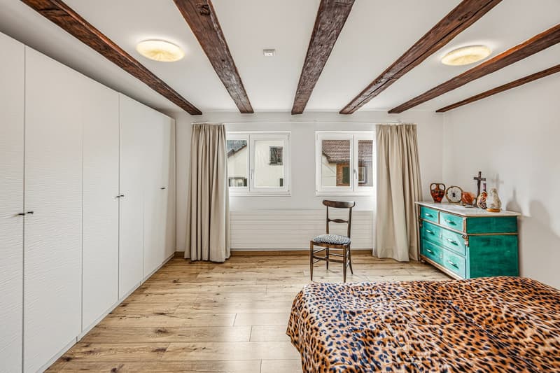 bedroom with wooden floor, white walls, wooden beams, wardrobe, bed with leopard print cover, chair, dresser, windows with curtains