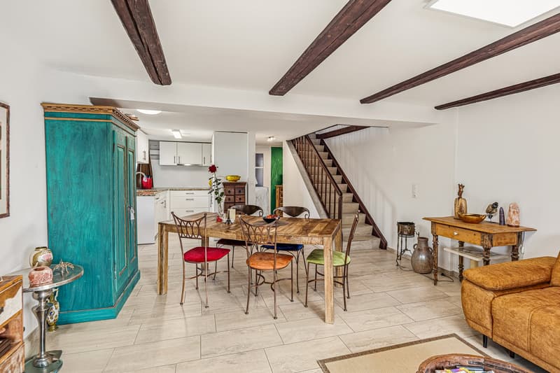 dining room with wooden table, chairs, blue cabinet, white walls, wooden beams, staircase, kitchen, couch, rug