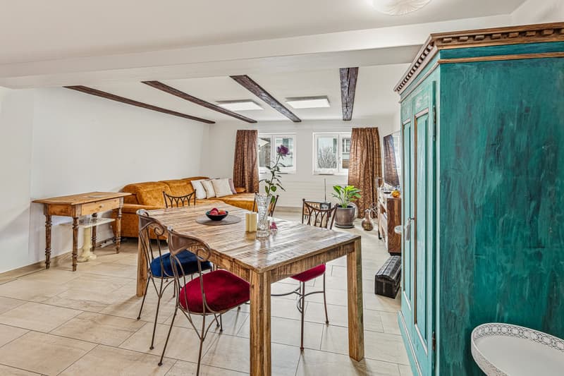 Dining room with wooden table, chairs, couch, wooden cabinet, potted plant, window with curtains, tiled floor