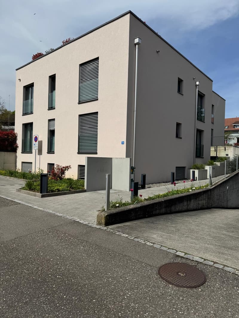 Modern apartment building with 4 floors, beige exterior, large windows, entrance with automatic gate, paved driveway, small garden area with plants and flowers, street sign, manhole cover