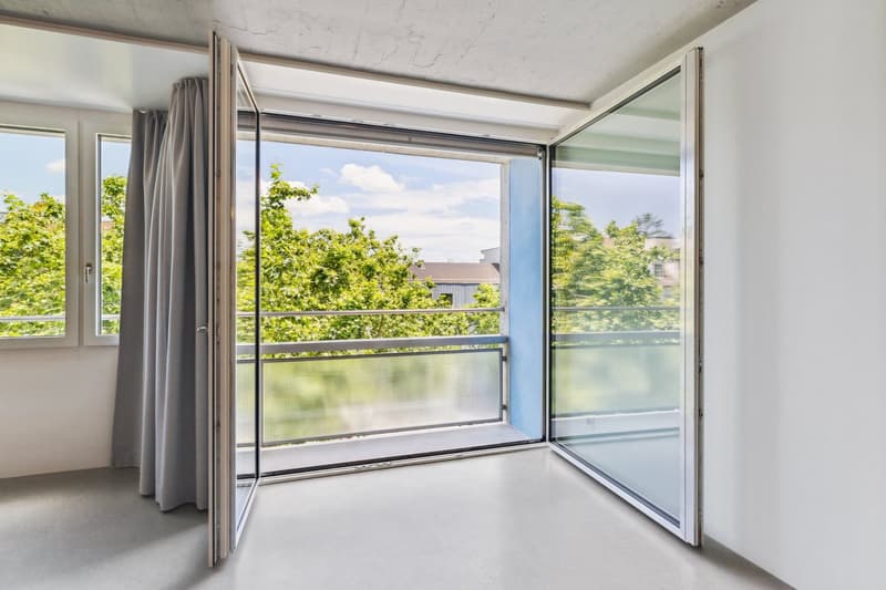Spacious living room with large windows, sliding glass doors leading to a balcony or terrace, and a view of the lush greenery outside.