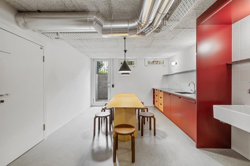 Open-concept kitchen and dining area with concrete floors, white walls, exposed ductwork, a large wooden dining table with stools, and a red kitchen cabinet unit with a built-in dishwasher.