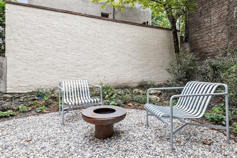 The image shows an outdoor seating area with two metal chairs and a round metal fire pit or fireplace in the center. The area is surrounded by gravel and greenery, with a tall white wall in the background.