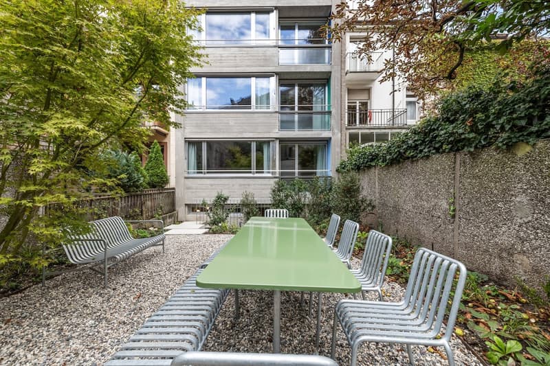 Multi-story concrete apartment building with large windows, surrounded by lush greenery and a paved outdoor seating area with a long green table and chairs