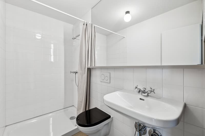 White tiled bathroom with a shower curtain, a sink with a faucet, and a toilet. The room has a clean, minimalist design.