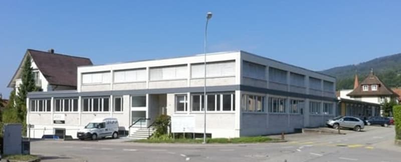A modern building with many windows, possibly an office or industrial building, cars parked in front, white walls, a van parked in front