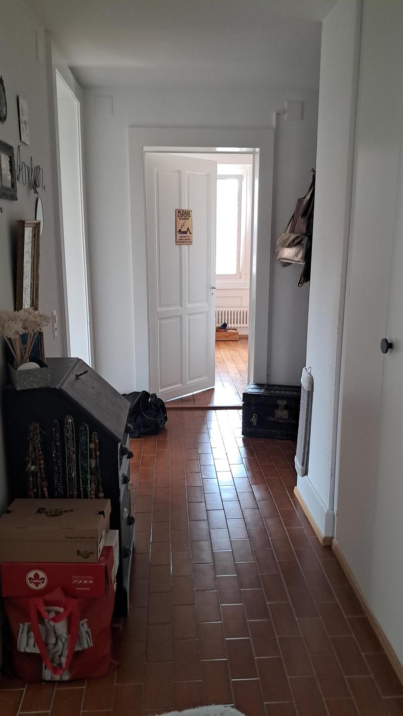 Empty hallway with white walls, brown tiled floor, open door, bags, boxes, and a sign