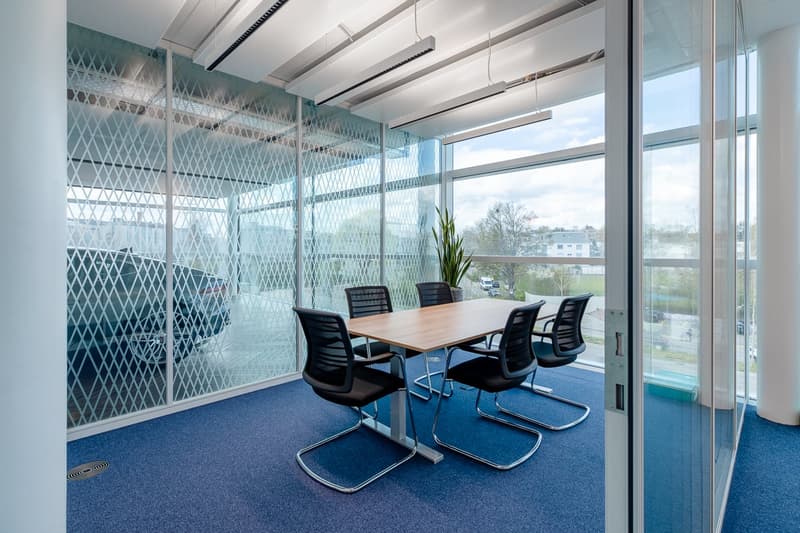 Spacious meeting room with large windows providing a view, wooden table, black chairs, glass walls with diamond pattern, and blue carpeted floor