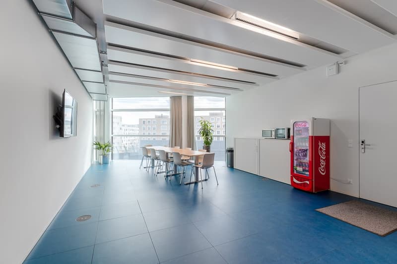 Large open office space with glass walls, exposed ceiling beams, dining table with chairs, and a Coca-Cola vending machine.