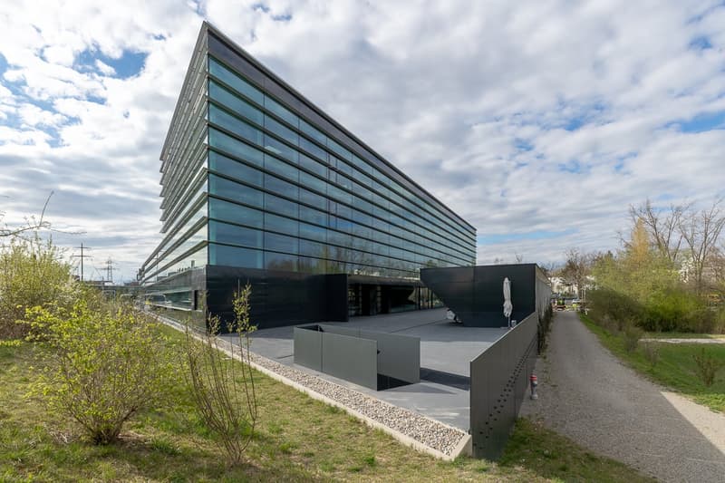 A modern, multi-story glass and steel building with a sleek, angular design. The building has a large, open entrance area and is surrounded by a grassy area with a paved walkway.