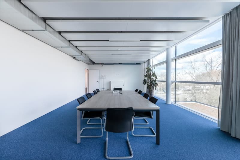 Large open office space with floor-to-ceiling windows, providing a view of the outdoors. The room has a long wooden conference table surrounded by black chairs, and a plant in the corner. The ceiling has exposed beams and lighting fixtures.