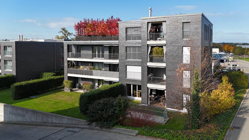 A modern, gray-bricked building with multiple floors, balconies on several levels, and greenery such as hedges and small trees.