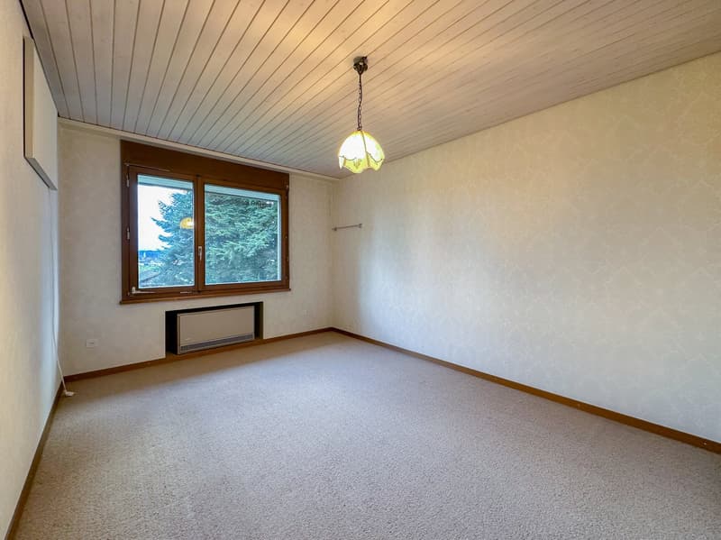 Empty room with beige carpet, white walls, wooden ceiling, large window, lamp hanging from ceiling