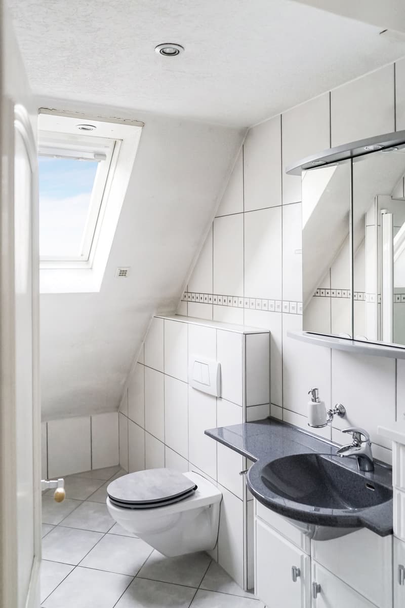 Modern bathroom, white tiles, grey countertop, white cabinet, toilet, sink, mirror, skylight