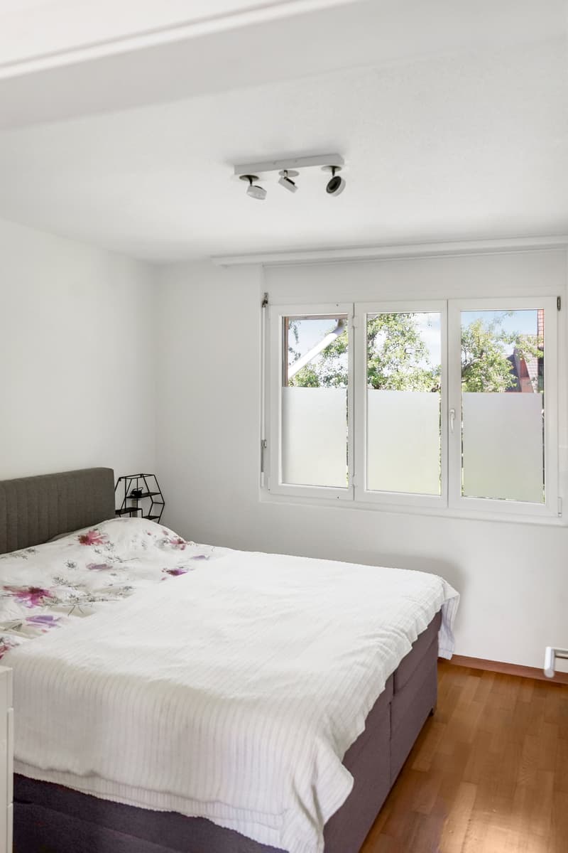 Spacious bedroom with large windows providing a view, white walls, hardwood floor, floral bedding, and a modern track lighting fixture on the ceiling.