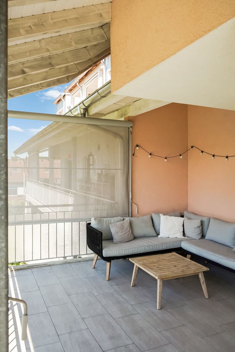 Covered balcony with gray tile flooring, a sectional sofa with gray cushions, a wooden coffee table, and string lights hanging overhead. The balcony overlooks a building exterior with a blue sky visible.