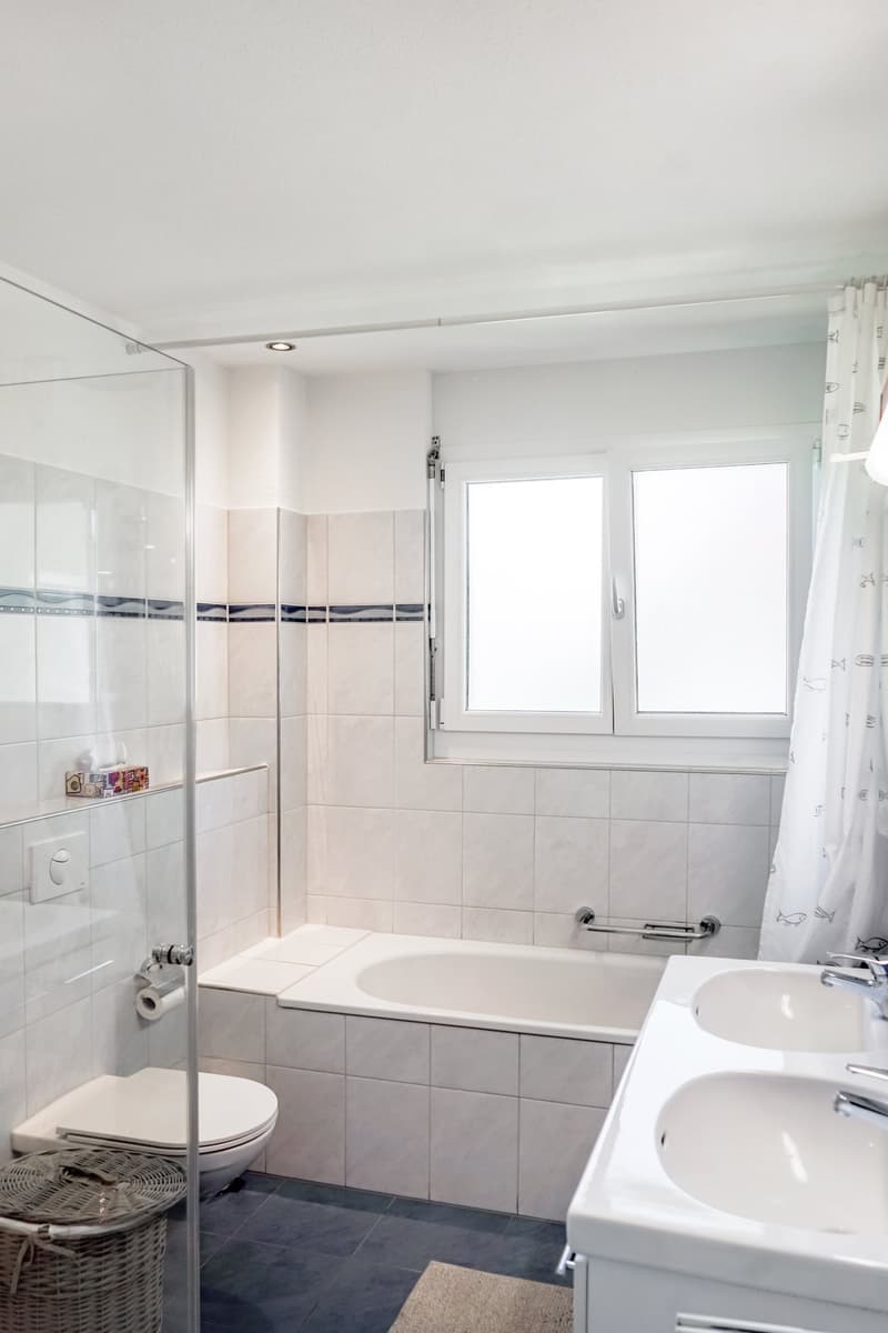 The image shows a spacious bathroom with a bathtub, a shower enclosure, and a vanity with a sink. The walls are tiled, and the floor is covered with dark tiles. The room has large windows that provide natural light. Overall, the bathroom appears to be cle