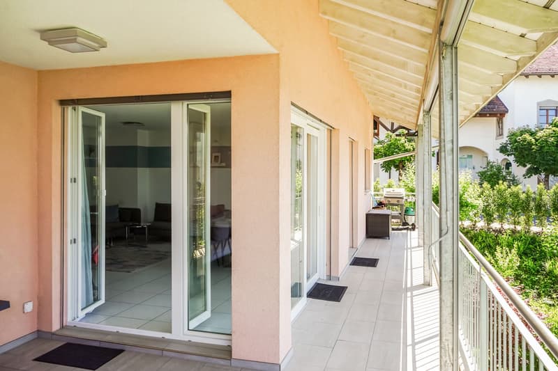 Spacious apartment with a balcony, large windows, and a covered walkway leading to the entrance. The interior is visible through the glass doors, showing a living room area. The exterior has a tiled floor and a railing along the balcony.