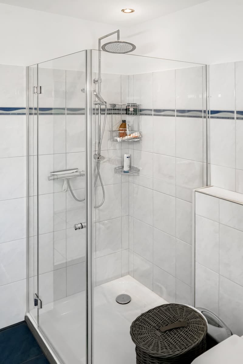 Modern bathroom with glass shower enclosure, tiled walls, shower head, and storage shelves.