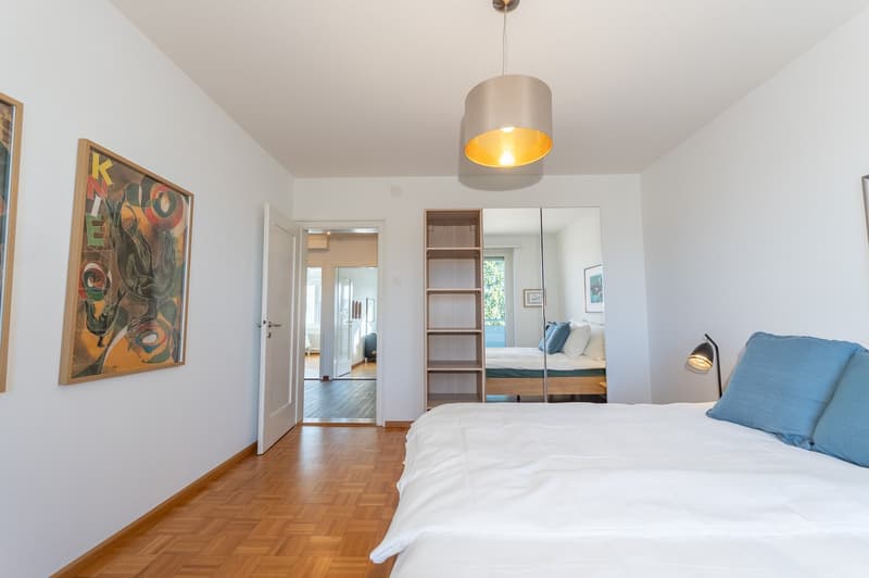 Large double bed, wooden floor, white walls, wall art, lamps, open door leading to another room, mirror reflecting another bed