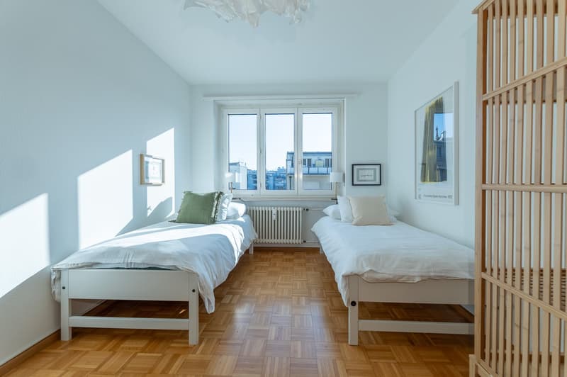 two beds with white sheets, pillow, wooden parquet floor, radiator, two picture frames, large window