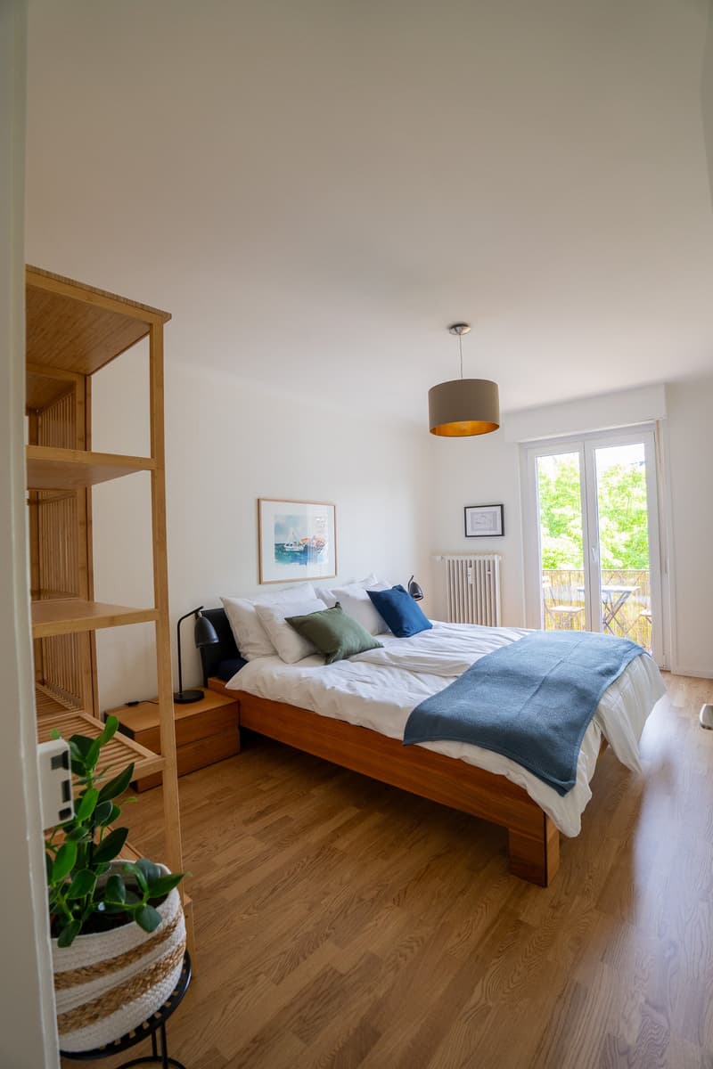 Spacious bedroom with hardwood flooring, white walls, large window leading to a balcony, wooden bed frame, bedside table, and a pendant light fixture.