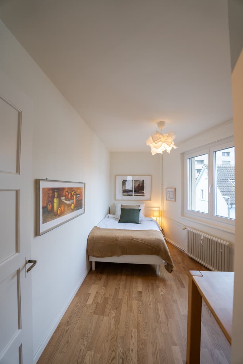 Spacious bedroom with hardwood flooring, white walls, large window, and a modern light fixture. The room features a bed with a green pillow, artwork on the walls, and a side table.