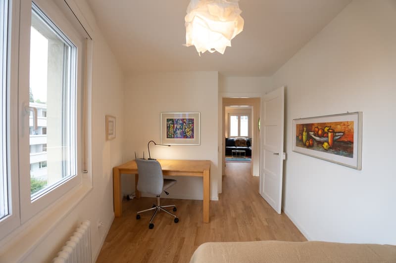 The image shows a bedroom with a wooden desk, a gray office chair, and a large window that provides natural light. The room has white walls and a hardwood floor. There are two framed artworks on the walls, and a decorative light fixture hangs from the cei