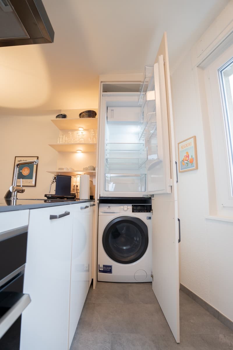 Compact kitchen with white cabinets, built-in dishwasher, washing machine, shelves, and coffee maker