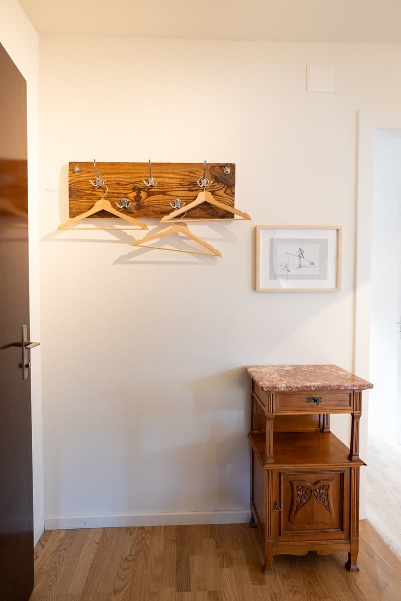 The image shows a room with a wooden wall-mounted coat rack, a wooden side table with a marble top, and a framed artwork on the wall. The room has a clean, minimalist aesthetic with white walls and hardwood floors.