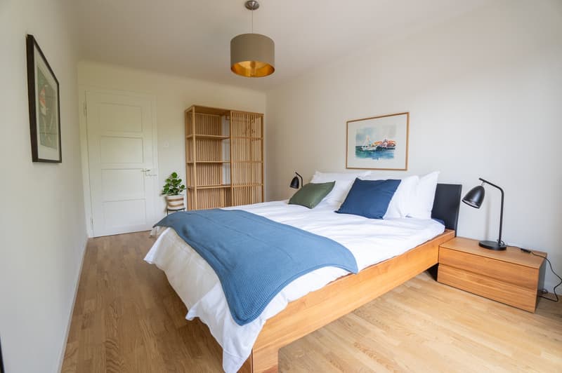 Spacious bedroom with hardwood flooring, white walls, large windows, wooden bed frame, bedside tables, and a bookshelf. The room has a cozy and modern feel with a blue and white color scheme.