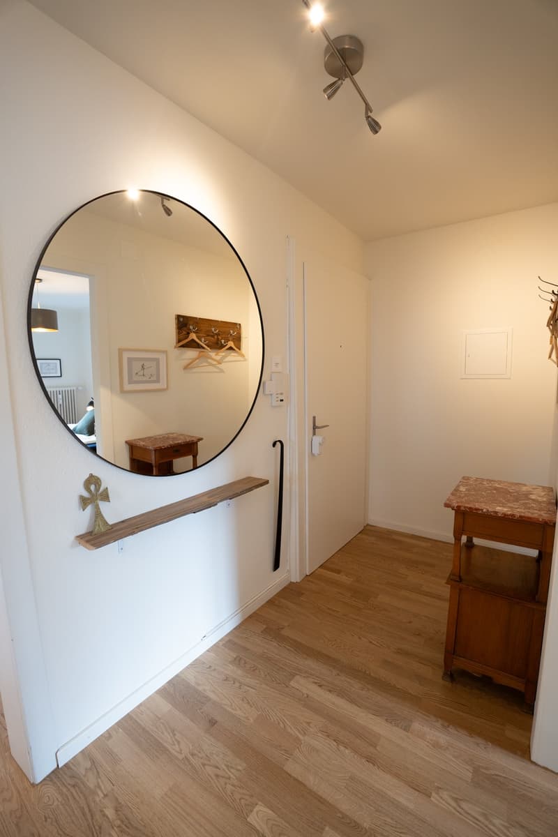 The image shows an entryway with a large round mirror on the wall, a wooden bench, and a wooden side table. The floor is made of hardwood and the walls are painted a light color. The lighting fixtures on the ceiling provide a warm, ambient lighting to the