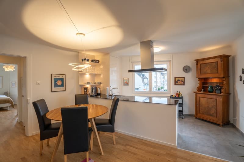 Open-concept kitchen and dining area with hardwood floors, white walls, and large windows. The kitchen features modern appliances including a dishwasher, and there is a round wooden dining table with black chairs. The room also includes a vintage-style ca